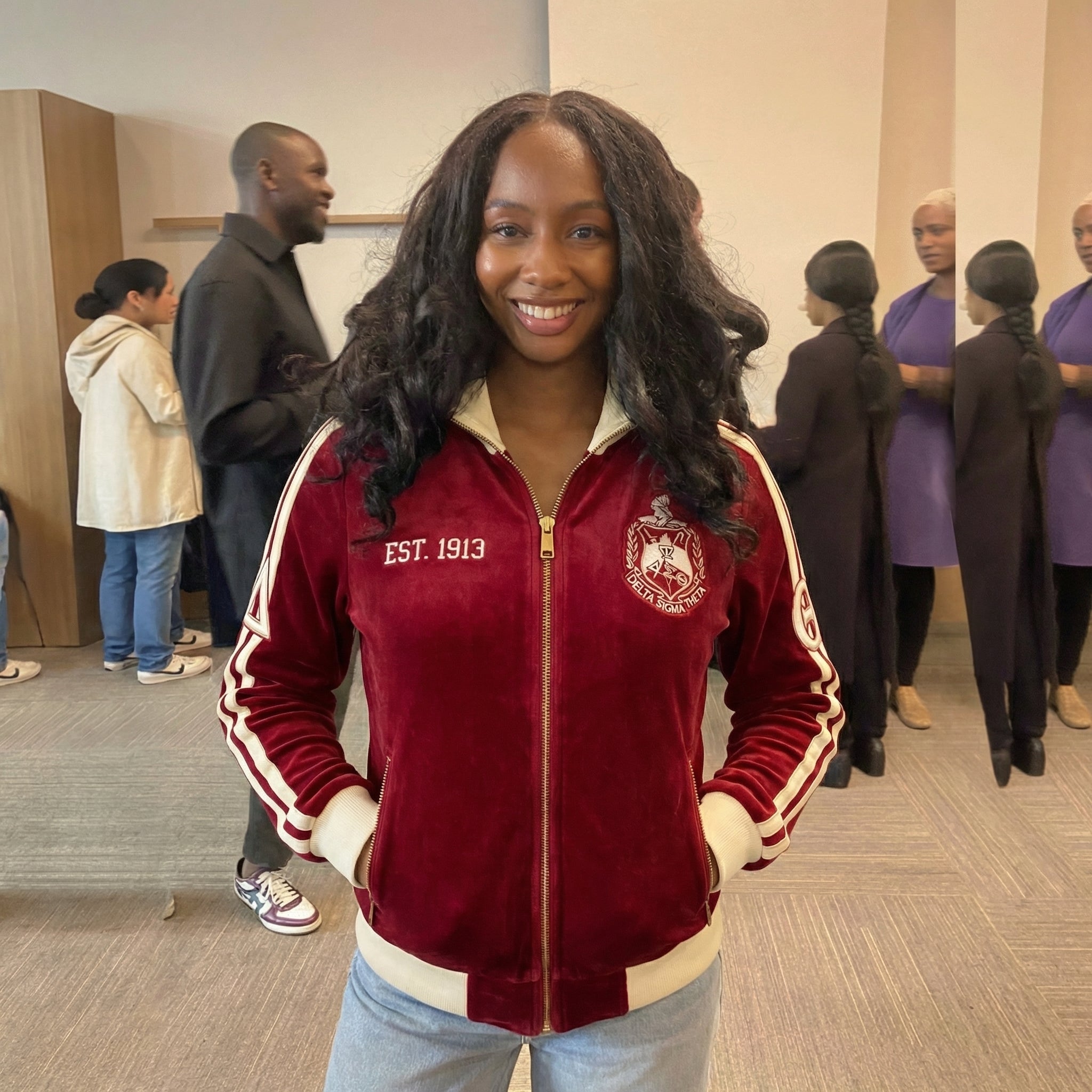Person wearing a red and white jacket with a logo, standing in an indoor setting with other people.