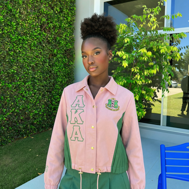 Woman wearing AKA Alpha Kappa Alpha pink green coach jacket with cursive script back pleated panels AKA letters and gold hardware styled outdoors
