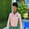 Woman wearing AKA Alpha Kappa Alpha pink green coach jacket with cursive script back pleated panels AKA letters and gold hardware styled outdoors
