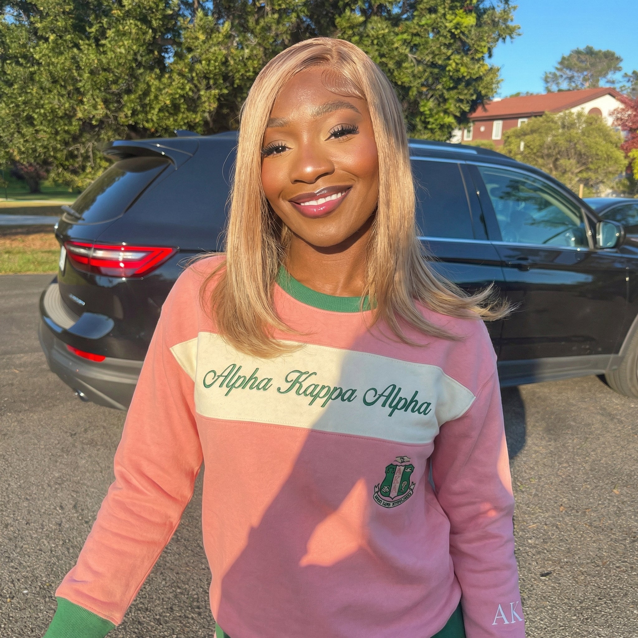 Woman wearing a pink sweatshirt with 'Alpha Kappa Alpha' text, standing in front of a black car.