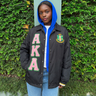 Person wearing a black jacket with 'AKA' letters and a crest, standing against a green leafy background.